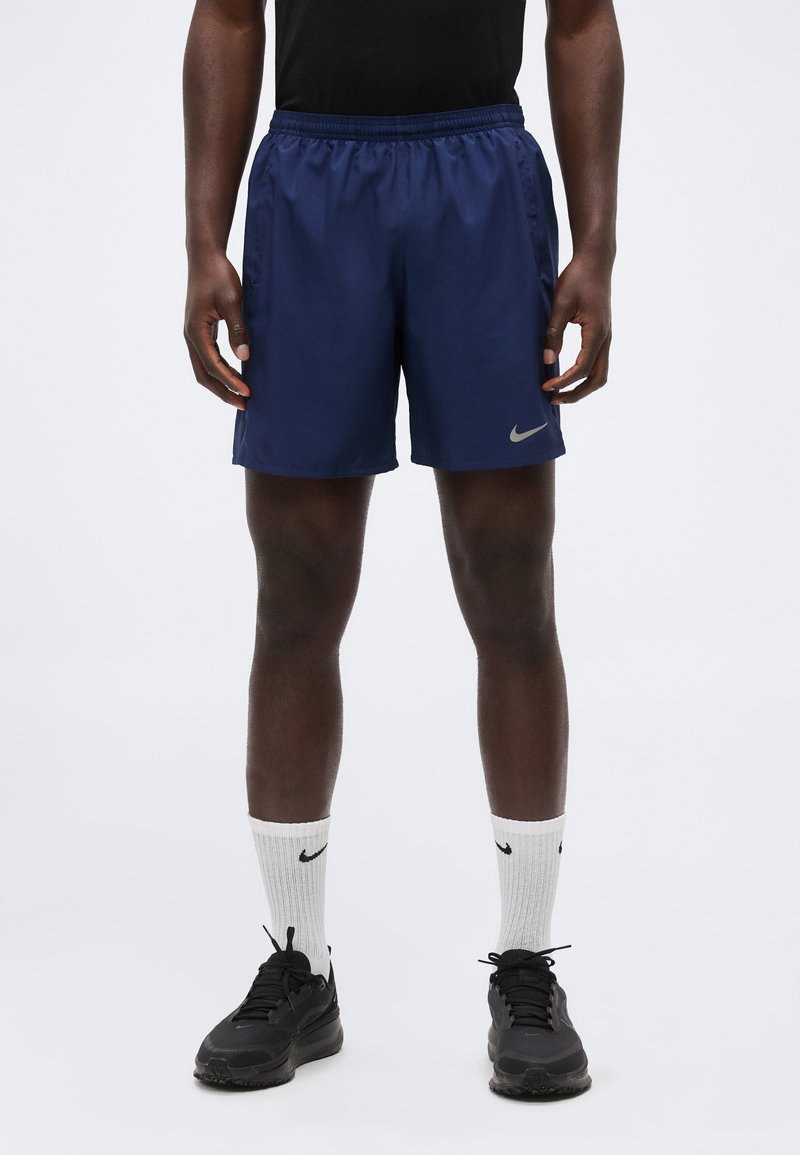 Άνδρας που φοράει μαρίν σορτς Nike, μαύρα αθλητικά παπούτσια και λευκές κάλτσες Nike, στέκεται μπροστά σε απλό λευκό φόντο.