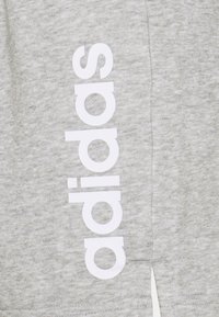 Grått tyg med vertikalt vitt "adidas"-logotyp tryckt på textilen, som visar en sidssöm och en liten tygslits.