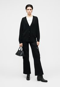 Cardigan noir à boutons, porté sur une chemise blanche. Assorti à un pantalon cargo noir et des bottines aux chevilles, ainsi qu'un petit sac à main noir.