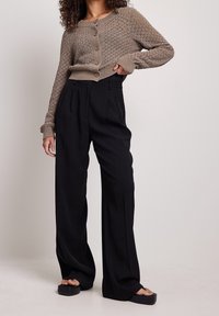 Ruskea neulottu neuletakki nappikiinnityksellä, yhdistettynä korkeavyötäröisiin mustiin leveälahkeisiin housuihin ja mustiin tasapohjaisiin sandaaleihin.