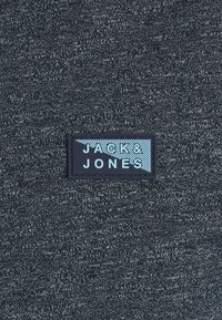 Grått textil med en strukturerad yta, med en svart rektangulär etikett med "JACK & JONES" i ljusblått, placerad centralt.