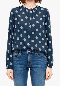 Long-sleeve navy blouse with white circular patterns, featuring a button front and gathered neckline, paired with blue jeans.