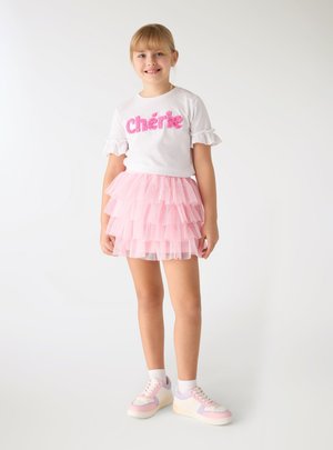 Młoda dziewczyna uśmiechająca się, ubrana w biały t-shirt z napisem „Chérie”, warstwową różową spódniczkę tutu, białe skarpetki oraz różowo-białe trampki, stojąca na białym tle.