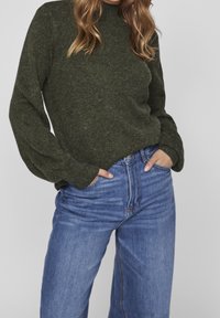 Mörkgrön stickad tröja med hög krage och oversized ärmar, kombinerad med ljusblå jeans med hög midja.