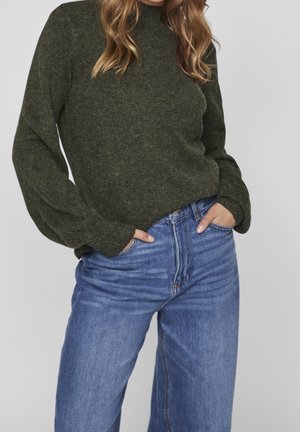 Maglione lavorato a maglia verde scuro con collo alto e maniche oversize, abbinato a jeans di denim azzurri a vita alta.