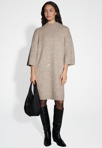 Abito oversized in maglia beige con collo alto e maniche a tre quarti. Abbinato a stivali neri in pelle alti fino al ginocchio e una borsa nera con paillettes.