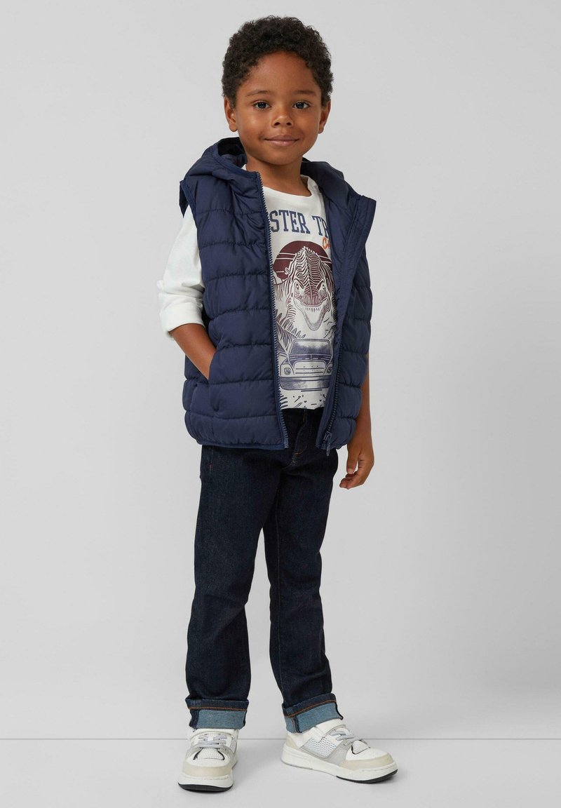 Marineblaue, gepolsterte Weste mit Kapuze, über einem weißen T-Shirt mit Grafik getragen. Dunkle Jeans und weiße Sneakers mit grauen Akzenten runden den Look ab.