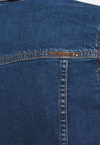 Zbliżenie na niebieską tkaninę dżinsową z pomarańczowymi szwami i małą metalową metką marki na górnym szwie.