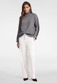 Szary sweter z golfem z wzorzystymi bocznymi akcentami, w połączeniu z białymi spodniami o szerokich nogawkach i brązowym obuwiem o fakturowanej powierzchni.