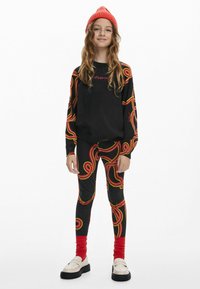 Schwarzer Sweatshirt mit bunten wirbelnden Mustern und Stickerei, passende Leggings mit ähnlichem Design, rote Socken und eine rote Strickmütze.