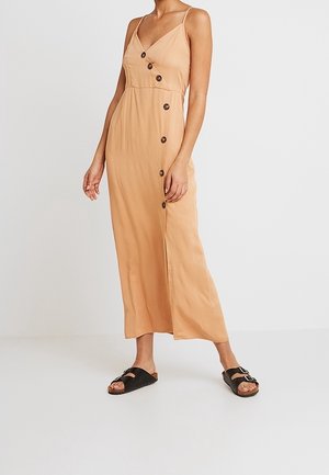 Vrouw draagt een mouwloze beige midi-jurk met knopen aan de zijkant en zwarte slip-on sandalen, staand tegen een effen witte achtergrond.