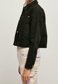 Svart kort denimjacka med knäppning och två bröstfickor; har en krage och subtilt texturerad tyg.