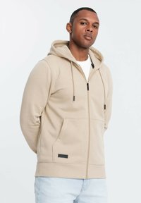 Sudadera con cremallera color beige, hecha de una tela suave, que cuenta con capucha, bolsillos frontales y cordones negros con puntas de metal. Diseño simple y funcional.