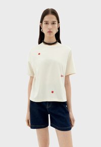 Crème korte mouwen t-shirt met rode bloemdessin, gecombineerd met donkere denim shorts. Heeft een ronde halslijn en een ontspannen pasvorm.