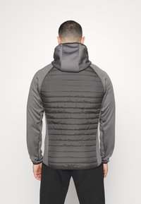 Grå huva jacka med en vadderad svart bakpanel och släta grå ärmar. Materialet verkar vara lätt men isolerat med en åtsittande passform.