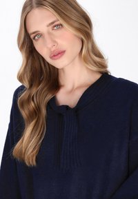 Pulover tricotat de culoare bleumarin cu un guler texturat și cravată, având o croială lejeră și mâneci lungi. Material moale, fără modele sau accesorii vizibile.