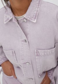Persona que lleva una chaqueta de mezclilla de color púrpura claro con botones y dos bolsillos en el pecho, con las manos descansando cerca de los bolsillos de la cintura.