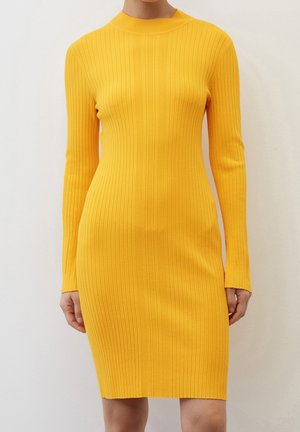 Femme portant une robe jaune moulante à manches longues côtelée qui arrive juste au-dessus des genoux, montrée du cou aux genoux.