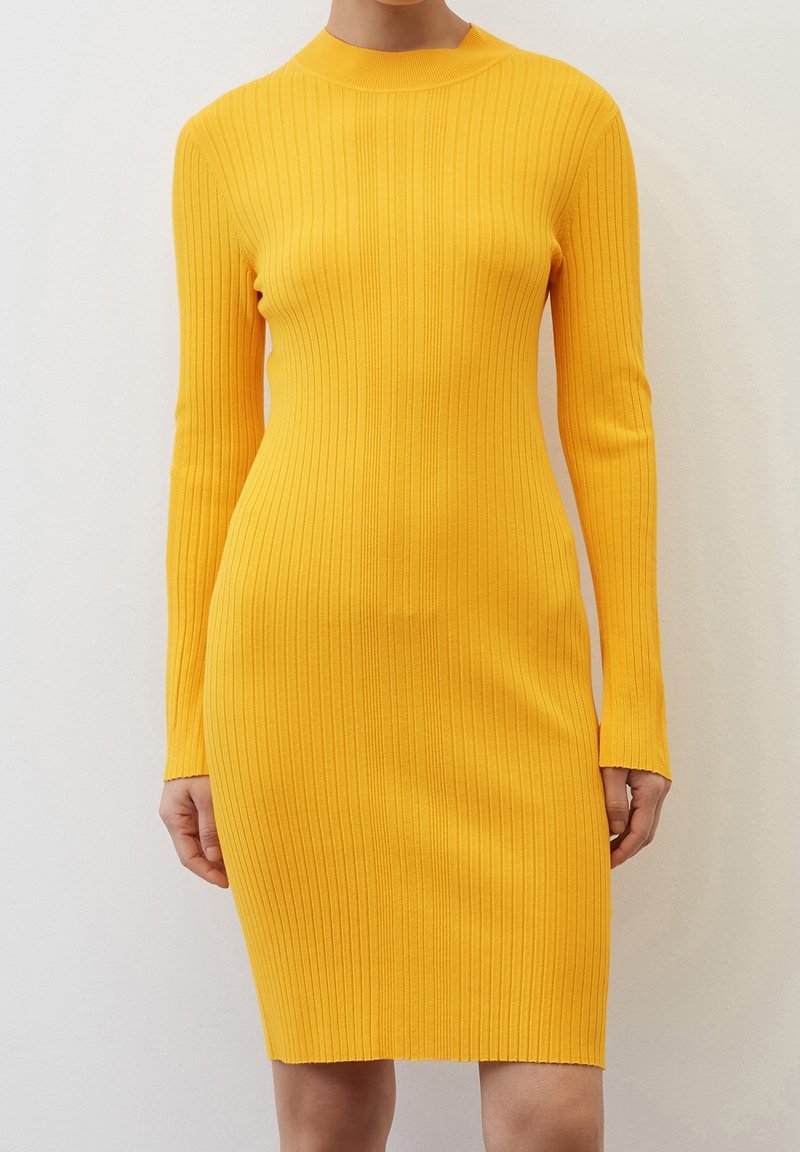 Femme portant une robe jaune moulante à manches longues côtelée qui arrive juste au-dessus des genoux, montrée du cou aux genoux.