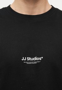 Κοντινό πλάνο στον λαιμό ενός μαύρου T-shirt με λευκό λογότυπο "JJ Studios" και μικρό κείμενο που αναφέρει την προέλευση του σχεδιασμού από τη Δανία.
