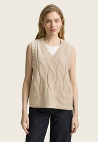 Gilet sans manches en tricot beige à col en V, présentant un motif de torsades texturé, une bordure côtelée et porté par-dessus un T-shirt blanc.