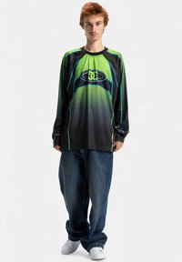 Camisola de manga comprida preta e verde com padrão em degradê e logótipo DC; usada com jeans azuis largos e ténis brancos.