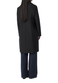 Cappotto lungo nero con taglio dritto, caratterizzato da un dettaglio della cucitura posteriore. Abbinato a jeans blu a gamba larga, mostrato da dietro.