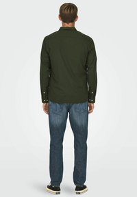 Camisa de manga larga verde oscuro con puños abotonados, combinada con jeans azul claro y zapatillas negras, vista desde atrás.