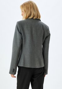 Blazer in misto lana grigia con una vestibilità sartoriale, colletto a scollo a incasso e polsini con bottone. Presenta un tessuto texturizzato e un design pulito e minimalista.