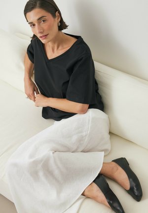 Zwarte katoenen V-hals shirt, witte linnen broek met wijdere pijpen en zwarte platte schoenen, zittend op een lichtgekleurde bank tegen een neutrale achtergrond.