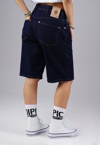 Person wearing dark blue denim knee-length shorts, white high-top sneakers, and white socks with black text. Hands in back pockets.