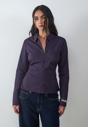 Woman with long dark hair wearing a fitted dark blue long-sleeve shirt with thin vertical stripes and dark blue jeans against a plain background.