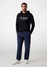 Sudadera negra con bordado "GUESS EST. 1981", combinada con pantalones de mezclilla azul oscuro y zapatillas blancas, presentada sobre un fondo neutro.