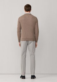 Sweter o splocie warkoczowym w jasnobrązowym kolorze, z fakturą prążkowaną, dopasowany krój, w zestawieniu z jasno-szarymi spodniami i ciemnymi butami. Widok z tyłu.