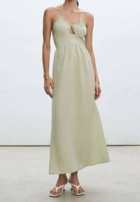 Robe maxi en tissu vert clair avec une découpe en trou de serrure, col halter, silhouette fluide et texture douce. Portée avec des sandales blanches.