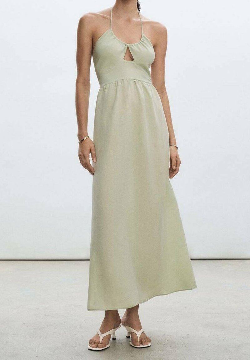 Robe maxi en tissu vert clair avec une découpe en trou de serrure, col halter, silhouette fluide et texture douce. Portée avec des sandales blanches.