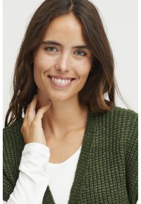 Olíva zöld, textúrázott mintázatú kötött kardigán, fehér, hosszú ujjú inggel párosítva. A modell keze gyengéden érinti a nyakát.