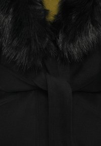 Melnā mētelī ar mīkstu, melnu mākslīgās kažokādas apkakli. Jostasvietā iekļauts sasienams jostas siksna, izgatavots no mīksta auduma ar gludu faktūru.