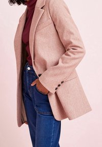 Růžový texturovaný blazer s jednou kapsou, výstřihy a černými knoflíky. Spárovaný s modrými džíny s vysokým pasem.