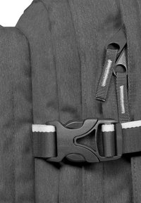 Details eines grauen Stoff-Rucksacks mit strukturiertem Material, schwarzem Kunststoffverschluss und schwarzem Reißverschluss mit weißen Akzenten.