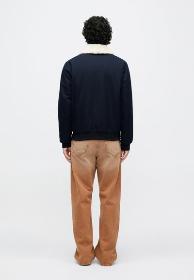 Modra jakna z ovratnikom iz umetnega krzna, nošena preko bež hlač. Jakna ima rebraste manšete in raven hrbet, kar poudarja preprost dizajn.