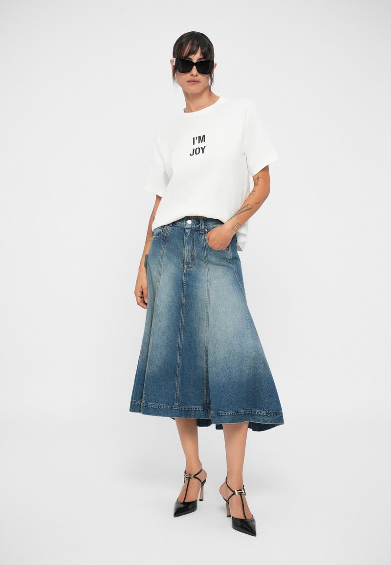Vit t-shirt med svart tryck "I'M JOY," kombinerad med en lång blå denimkjol. Modellen bär svarta solglasögon och svarta klackskor.