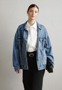 Chaqueta de mezclilla azul de gran tamaño con dos bolsillos en el pecho, llevada sobre una camisa blanca de botones, combinada con pantalones negros de corte ajustado y un bolso de mano.