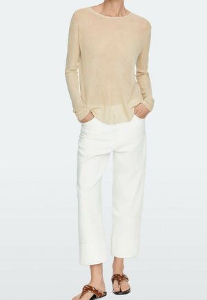 Personne portant une chemise beige à manches longues, un pantalon blanc large raccourci et des sandales marron à motif animalier, debout les mains dans les poches.