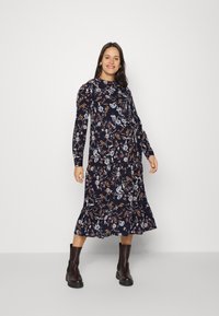 Vestido floral de manga longa em azul marinho com um acabamento em babados, apresentando uma mistura de flores brancas, pink e amarelas. Usado com botas de tornozelo escuras.