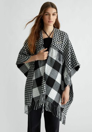 Poncho a scacchi bianco e nero con bordi frangiati e un contrastante motivo houndstooth. Realizzato in tessuto morbido, si drappeggia liberamente sul corpo.