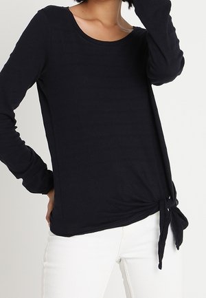 Kobieta ubrana w czarną bluzkę z długim rękawem z wiązaniem po boku, w połączeniu z białymi spodniami, stojąca na tle o jednolitym kolorze.