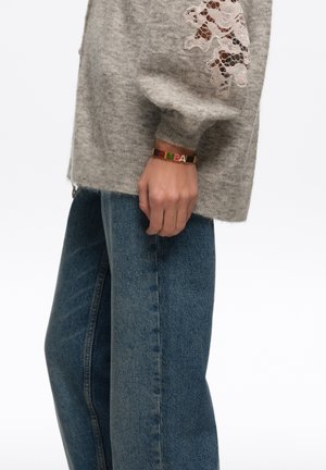 Persona vestida con vaqueros azules y un suéter gris con detalles de encaje, mostrando una pulsera dorada con dijes de letras coloridas en su muñeca.