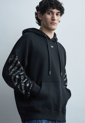 Młody mężczyzna o kręconych włosach, ubrany w czarną bluzę z kapturem z wzorami srebrnych łańcuchów na rękawach oraz małym napisem "OFF" na piersi, z rękami w kieszeniach.