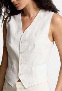 Bezrukávý bílý polokošilový vest s výstřihem do V, nosený přes odpovídající kalhoty světlé barvy, s detailním záběrem na horní část trupu.